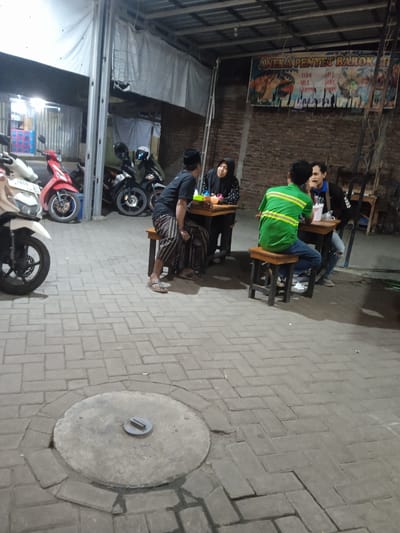 This image depicts a group of people gathered at an outdoor food stall or restaurant, likely in Cangkring, Indonesia. Four individuals are visible, seated around small wooden tables. One man, wearing a black shirt and a peci hat, sits facing away from the camera, his legs crossed. To his left, a woman in a black hijab is seated at another table, looking towards the man. In the foreground, a person in a bright green shirt with yellow reflective stripes is seated on a stool, also facing away from the camera. Beside them, another man in a blue shirt is visible, looking towards the person in the green shirt.
The setting appears to be an open-air establishment with a corrugated metal roof supported by metal beams. The floor is paved with interlocking grey bricks. Several motorcycles are parked in the background, including a white scooter on the left and two black scooters to the right. The time of day is likely evening or night, as indicated by the artificial lighting and the shadows cast on the ground.
A banner hangs on the brick wall behind the tables, featuring colorful imagery and text. The visible text on the banner includes "ANEKA PENYET BAROKAH" and lists food items such as "AYAM," "LELE," "NILA," and "GURAME." The overall atmosphere seems casual and relaxed, with the patrons engaged in conversation or enjoying their meal. The presence of multiple motorcycles suggests it might be a popular local spot.