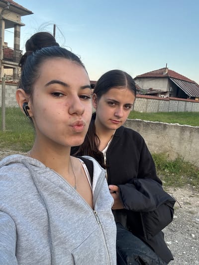 Two young women are in the foreground of an outdoor, daylight scene. The woman closest to the camera is wearing a gray hooded sweatshirt, a black wireless earpiece in her left ear, and a silver necklace. She has dark hair pulled back in a bun and is making a kissing face. The second woman, partially visible behind her, wears a black jacket over a white top.
In the background is a combination of urban and rural elements. A concrete wall and a building with a red-tiled roof are visible. The sky is a pale blue. Given the prompt's specification, the location is likely Karavelovo, Bulgaria.