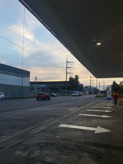 The image depicts a view from what appears to be a gas station in Quilmes, Argentina. An orange-clad individual is seen near the station's price display. The camera faces a street with visible traffic. Road markings include large white directional arrows. Power lines are visible against the sky, with buildings lining the street. Cars are parked along the street. The sky is light, suggesting either dawn or dusk.