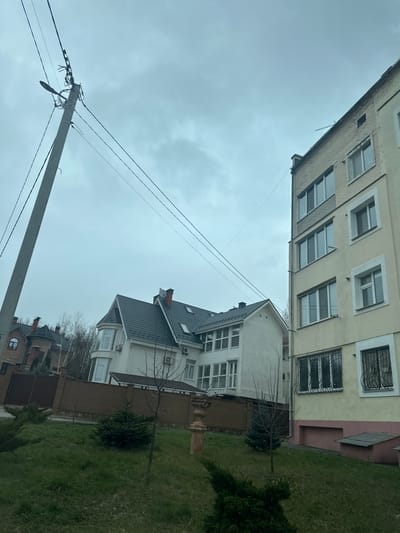 The image shows a street scene in Yurivka, Ukraine (latitude: 50.332, longitude: 30.372). In the foreground, there is a brick fence, a grassy lawn, and a few evergreen trees. A concrete utility pole with several electrical wires running across the frame stands on the left side of the image.  To the right, a tall, three-story, residential building with a beige facade and several windows is visible. The windows are mostly rectangular, with a few square ones. The building has a red brick base, a small red brick overhang above the windows on the first floor, and some black metal bars in front of the windows on the first floor. In the center and background, a residential building with a gray roof, white walls, and several windows is partially visible. The building has a decorative wooden door and some shrubs in front of it. The sky is overcast and gray.