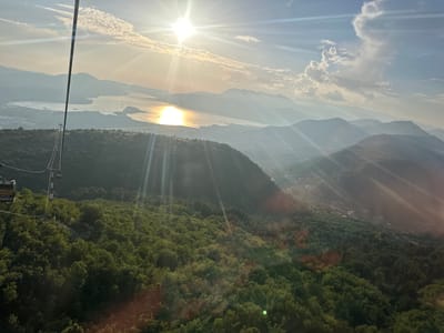 The image depicts a panoramic view from a ski lift or cable car descending over a lush, forested mountainside towards a serene body of water. The sun is setting or rising, casting a brilliant golden glare across the water and creating long, ethereal light rays that pierce through the hazy atmosphere. The water, possibly a bay or lake, is dotted with a few small boats, and a coastal town or village is visible along its edge, nestled between the water and the rolling hills. Further inland, multiple layers of mountains recede into the distance, their peaks softened by the atmospheric haze. The foreground is dominated by the dense green canopy of trees, with the cable car's support tower and a section of the cable visible in the upper left quadrant. The ski lift appears to be traveling from the mountains towards the water, offering a breathtaking perspective of the natural landscape. The overall scene conveys a sense of peacefulness and natural grandeur, characteristic of the mountainous coastal regions of Montenegro, specifically the area around Mirac.