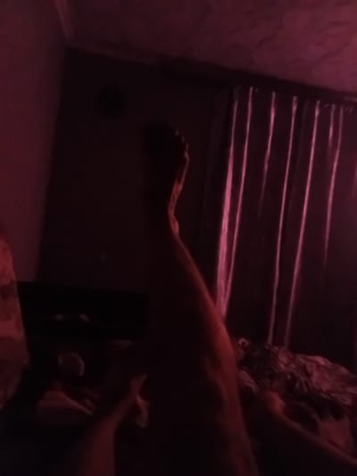 The image depicts a dimly lit bedroom interior, potentially in Verkhniachka, Ukraine, under artificial red lighting. In the foreground, a person's leg is visible, extending from the bottom left of the frame upwards and to the right. The person appears to be lying down or reclining. Behind the leg, a patterned fabric, possibly bedding or a blanket, is visible. Further in the background, draped curtains in a dark color with subtle vertical folds are present on the right side of the frame. The overall ambiance is dark and suggestive, with limited clear details due to the low light conditions and color cast.