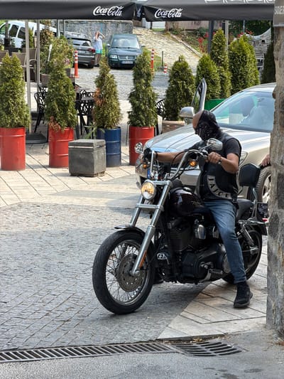 A person is sitting on a motorcycle parked on a cobblestone street. The person is wearing a black vest with "JAJCE" written in white letters, blue jeans, and a black bandana. The motorcycle is black with chrome accents. In the background, there is a white car, several potted evergreen trees, and some outdoor seating with tables and chairs. A black Coca-Cola umbrella is partially visible on the left side of the frame. The scene appears to be outdoors, possibly in a town square or cafe area. There is a metal grate on the right side of the street. The lighting suggests it is daytime, and the weather appears to be clear. The location is identified as Jajce, Bosnia and Herzegovina.