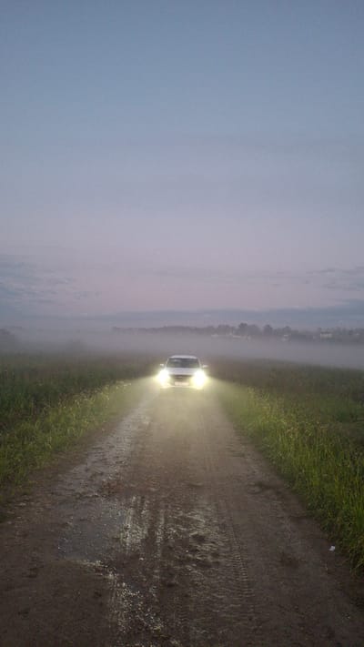A white car with its headlights on drives down a muddy dirt road in Kaporskoe, Russia, at dusk. The car is facing the camera, and its headlights illuminate the road and the surrounding fog. The road is wet and reflects the headlights. On either side of the road are grassy fields. In the distance, silhouetted trees and buildings can be seen through the fog. The sky is a pale blue and gray, indicating the transition from day to night.