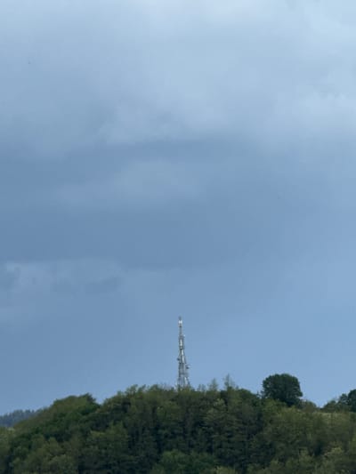 The image shows a daytime view of a cloudy sky above a forested area. A communication tower is visible amongst the trees. The location is identified as Zhaltusha, Bulgaria.