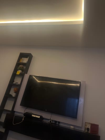 A television is mounted on a wall, with a soundbar beneath it. To the left of the television is a tall, narrow bookshelf with several items on it, including decorative plants and what appears to be a toy car. The room is lit by a recessed ceiling light that emits a warm glow. The walls are painted a light color, and the overall impression is one of a modern, uncluttered living space. The image is taken from a low angle, looking upwards towards the ceiling. No people are visible in the image. This scene is located in Nabatum, Nigeria.