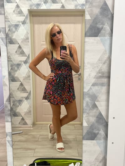 A woman with blonde hair is taking a selfie in a mirror. She is wearing sunglasses and a colorful dress. She is standing in what appears to be a room with geometric wallpaper and wooden flooring. The mirror reflects the woman, a door, and part of the room. She is holding a phone in her right hand, which is out of frame. The overall mood is casual and relaxed. There is no visible text in the image. The location is Ivanovskaya, Russia.