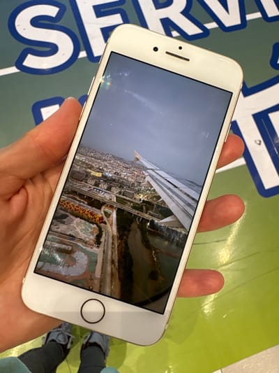 A human hand, appearing light-skinned, holds a white smartphone vertically over a glossy, lime-green surface. The phone's screen displays an aerial photograph, viewed from an aircraft. The image on the screen depicts a partial white aircraft wing with visible flaps extending over a sprawling urban landscape. This landscape features numerous buildings, an extensive network of roads and bridges, a winding river or canal, and a distinctive multi-colored (yellow and red) patterned area. The sky above the urban area is overcast and dim. Below the hand and phone, the lower legs of an individual wearing light blue trousers and grey-and-white sneakers are visible. The green background surface bears large, partial white and dark blue lettering, spelling out "SERV," likely part of a word like "SERVICE." The depicted urban landscape is located in Tianya District, China.