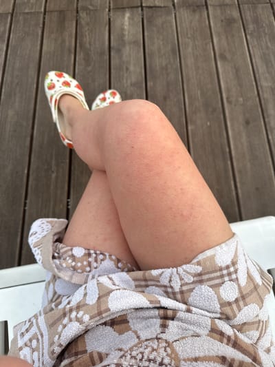 The image shows a close-up view of a person's legs, with their feet resting on a wooden deck. The person is wearing white slip-on shoes with a strawberry pattern. Their legs are crossed, and a towel with a floral and plaid pattern is draped over their lower body. The background is a wooden deck, and the lighting suggests it is daytime. The location is Yuzhnyye Koryaki, Russia.