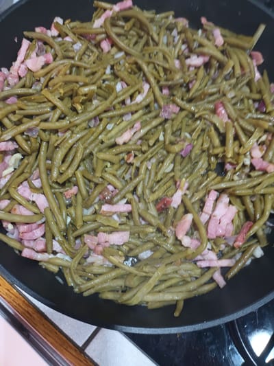A close-up shot displays cooked green beans with bacon in a black frying pan, situated on a stovetop. The beans appear to be slightly overcooked, with a brownish-green hue. The bacon is cut into small, rectangular pieces, scattered throughout the dish. There are also some diced onions visible. The pan is resting on a ceramic stovetop, partially visible. The image was presumably taken in Aubange, Belgium.