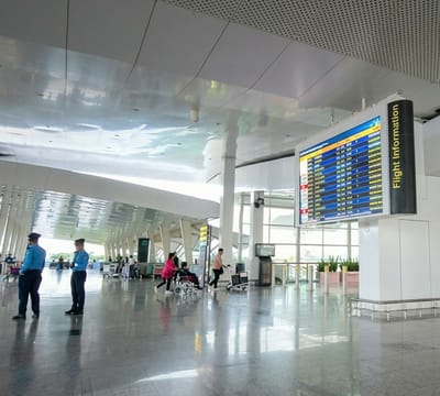The image captures a spacious and modern airport terminal, likely during the daytime due to abundant natural light streaming in from large windows visible in the background. The scene is dominated by a clean, highly reflective floor and a high, intricately designed ceiling with both smooth, reflective panels and perforated sections.
In the foreground, two security guards in blue uniforms and hats stand facing away from the camera. Further into the terminal, several individuals are present, including a person in a pink top pushing another person in a wheelchair. Another individual is seen pushing a luggage cart. In the background, more people are visible, some with luggage carts, suggesting typical airport transit activity.
A prominent feature is a large, rectangular digital flight information display hanging from the ceiling on the right side of the frame. It shows multiple rows and columns of flight data, including what appears to be flight numbers, destinations, and times. Vertically aligned on the right edge of this display, the words "Flight Information" are clearly visible against a yellow background.
The overall setting is one of organized movement and waiting. The architecture is contemporary, with tall, slender columns and a sense of openness. Greenery in planters can be seen near the windows in the mid-ground, adding a touch of nature to the sterile environment. The reflections on the floor and ceiling create interesting visual effects, mirroring the light and structure of the terminal. The general atmosphere appears calm and orderly.