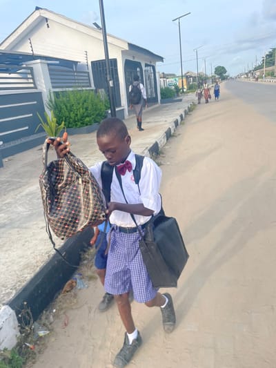 An outdoor scene in Mosafejaw, Nigeria, depicts a group of students on a dirt and paved roadside. In the foreground, a male student, appearing to be of elementary school age, is dressed in a white short-sleeved collared shirt with a red bow tie, blue and white checkered shorts, and dark sneakers with white socks. He carries a black shoulder bag on his left side and holds a patterned, glossy black and gold drawstring backpack in both hands, looking down at it. His expression is focused downwards. Behind him, another male student is partially visible, wearing blue shorts and dark shoes.
To the student's left, a paved sidewalk with a black and white striped curb runs alongside the scene. Beyond the sidewalk, a white and gray building with a gated fence and security wire is visible. Green shrubs grow in front of the building. Further down the sidewalk, another student wearing a gray uniform and a black backpack walks away from the camera.
In the mid-ground and background, a wide, unpaved road stretches into the distance. Several other students, predominantly female, wearing various school uniforms and carrying bags, walk along the right side of the road away from the camera. Utility poles with streetlights line the road. The sky is clear with scattered clouds. The overall lighting suggests daytime.