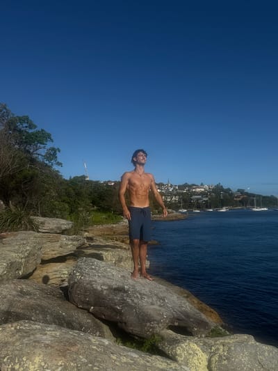An outdoor, daytime scene in Vaucluse, Australia, depicts a single male subject standing on a large, grey rock outcrop. The subject, a barefoot man with dark hair, is shirtless, wearing dark blue swim shorts, and a pendant necklace. He stands with his head tilted upwards, eyes closed, and arms relaxed at his sides, facing towards the right. To the subject's right and extending into the middle ground is a calm, dark blue body of water, likely a bay, dotted with numerous white sailboats and yachts at anchor. The distant shoreline features a green hillside heavily vegetated with trees and bushes, upon which various residential buildings, including a structure with a visible spire, are constructed. A construction crane is visible above the tree line on the left side of the distant hill. The sky is clear and bright blue, devoid of clouds. The foreground rocks display varied textures and shades of grey and beige.