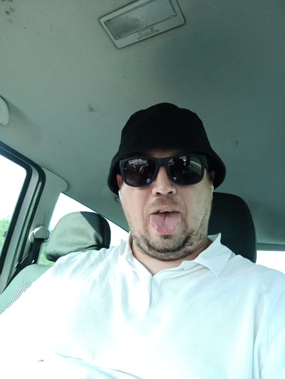 A man is taking a selfie inside a car. He is wearing a black bucket hat and sunglasses, and has his tongue sticking out. The interior of the car is visible, including the ceiling light and a headrest. The man is wearing a white collared shirt. The scene appears to be during the daytime, as indicated by the bright light coming from outside the car. The location is Všechromy, Czechia. No visible text is present in the image.
