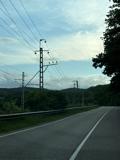 The image shows a road with power lines running alongside it. The road is asphalt and has a white line marking the edge. To the left of the road, there is a grassy verge, followed by dense green trees. In the background, there are more trees and a hill. The sky is partly cloudy, with some bright white clouds against a pale blue background. The sun appears to be setting or rising, casting a soft light. The power lines are supported by metal poles, and several wires are visible, stretching across the frame. The scene appears to be a rural or semi-rural area, possibly a highway or a major road passing through a forested landscape. There is no text visible in the image. The overall impression is one of a tranquil, natural setting with man-made infrastructure.