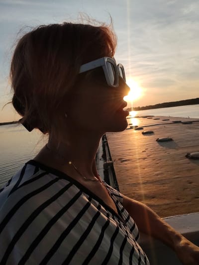 A mid-shot, profile portrait of a woman with reddish-brown hair, wearing white-framed sunglasses and a black and white striped shirt, is captured against a sunset sky. The sun is positioned directly behind her head, creating a strong lens flare and silhouetting her features. She is looking towards the right of the frame. In the background, a wooden pier extends into a body of water, reflecting the warm hues of the sunset. The pier appears to be located in the city of Shchetinino, Russia. The overall scene evokes a sense of tranquility and a connection with nature.