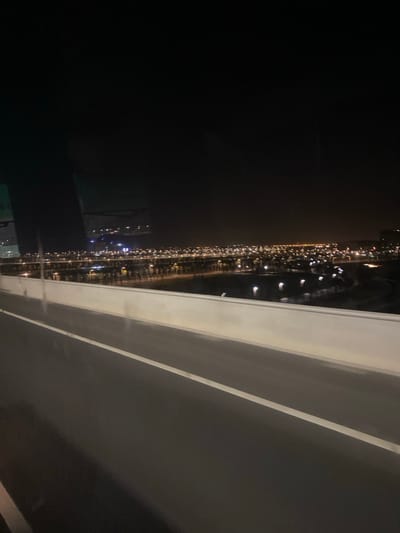 An aerial night view reveals a dark expanse punctuated by a distant, linear string of lights suggesting a city or road. A solid, light-colored structure dominates the foreground, potentially a balcony or edge of a building. Its stark contrast with the dark sky emphasizes the height from which the photograph is taken. The overall impression is one of urban observation during nighttime.