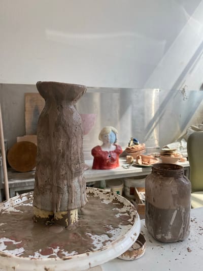 The image shows a ceramic studio, possibly in Saint-Ouen-sur-Seine, France. A clay sculpture on a dirty white wheel is the primary subject. The sculpture appears unfinished, with a roughly shaped body and head. Behind it, there is a painted clay bust of a woman, dressed in a red top and with light hair. Other clay figures and pottery are also visible in the background, including a dirty glass jar on a white surface. The surfaces in the room show signs of clay residue. The lighting is bright and appears to be natural. The background includes a metallic wall or shelf.