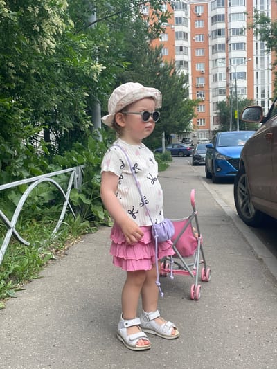 A young girl wearing a pink hat, sunglasses, and a pink and white shirt is standing on the sidewalk in front of a large apartment building. The girl is wearing a pink skirt and sandals. She is holding a purple purse and pushing a pink doll stroller. The stroller is in the background on the right side of the image. There are several other cars parked on the street in the background. The location is Москва, Russia, based on the coordinates provided. The scene is on a city street, likely a residential area. The sidewalk is gray and the building is beige brick. The girl is facing forward and is looking towards the right. The image was taken at an angle.