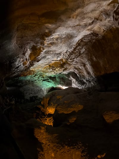 The image shows a dimly lit cave interior in Haría, Spain. The cave walls are rough, textured, and vary in color from light gray to dark brown. A narrow opening lets in a green-tinted light, illuminating a portion of the cave. The floor is dark and rocky. The image contains no visible people, and the focus is on the geological features of the cave.