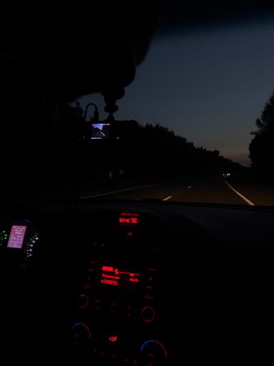 This image, taken at dusk or night on a highway near Vargany, Russia, provides a view from inside a car. The dashboard displays illuminated information. On the left, a digital instrument cluster shows a speed of approximately 60 km/h, 486 km remaining, fuel economy of 7.2 L/100, and a mileage of 205819 km. The gear is in 'D' for drive. To the right, a car stereo system emits a red glow, displaying the time as 21:00:55 and a radio frequency of 103.70. The car's internal temperature is displayed as 25°C. The road ahead is visible with lane markings and the headlights of other vehicles in the distance. Trees line the sides of the highway, and the sky is a deep twilight blue. A dashcam is mounted on the windshield, and its screen shows a road view.