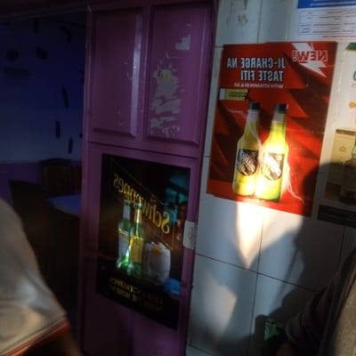 The image shows the interior of a bar or shop in Kinoo ward, Kenya. A purple cupboard with two posters is visible. One poster is a "Schweppes" advertisement and the other is for "New! Taste it! Charge it". The second poster contains two bottles of a yellow-green drink. There is white tiling on the wall next to the cupboard and a partial view of a person's body in the foreground.