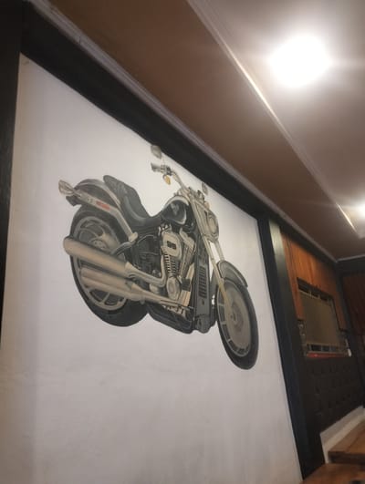 The image is a photograph of a mural of a motorcycle painted on a white wall. The motorcycle is depicted in profile, with its front wheel on the left and its rear wheel on the right. The motorcycle is black and silver, with chrome accents. It has a large engine, a comfortable seat, and a sleek design. The mural is highly detailed and realistic, with intricate shading and texture. The lighting in the image suggests an indoor setting, possibly a restaurant or a bar, with a warm, ambient glow coming from overhead lights. The background features a brown wooden ceiling and part of a wall clad in dark material with wood paneling above. The context of Bandarbireuen, Indonesia, is not visually present in the provided image.