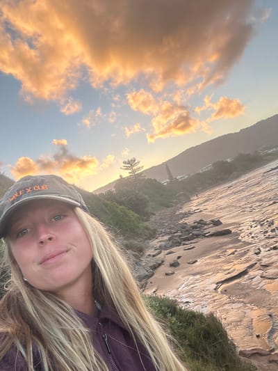 A medium-close-up shot of a woman with blonde hair and a baseball cap, seemingly taking a selfie, in front of a coastal landscape. The sky is visible with orange-tinted clouds, suggesting either sunrise or sunset. Part of a rocky cliff and coastline is visible in the background. Wollongong City Council, Australia, would be the location based on the coastal landscape and information provided.
