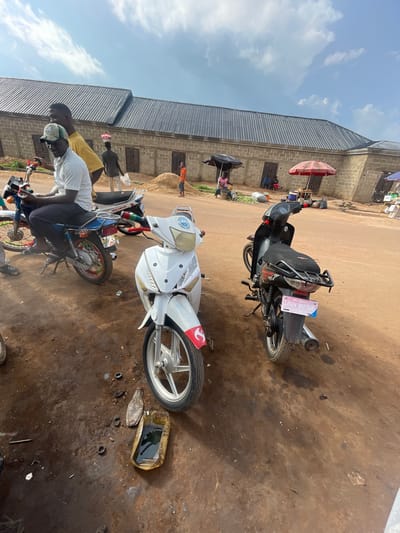 Two motorcycles are parked on a dirt ground in front of a building with a metal roof. One motorcycle is black with a pink license plate that reads "LIFAN MOTOR". The other motorcycle is white with red accents. There are a few people present, some standing and some sitting on the motorcycles. A red umbrella is set up near the building. The scene appears to be an outdoor market or workshop. The time of day appears to be during daylight hours. The weather seems to be clear with some clouds in the sky.