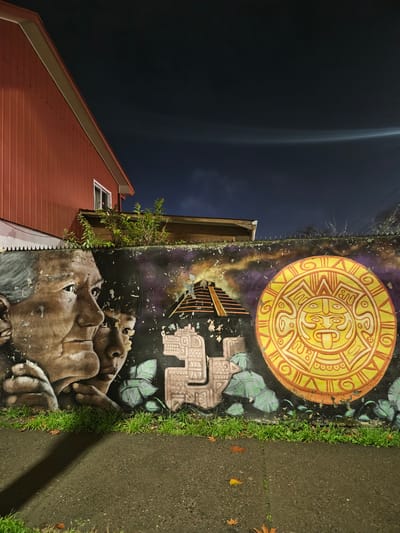 A night-time street-level shot captures a mural on a concrete wall in Chillán, Chile. The mural features a stylized depiction of two faces, an elder and a child, rendered in shades of brown and gray. To the right of the faces is a bright yellow circular motif resembling a sun or calendar stone, with intricate patterns and a central face with its tongue extended. Behind these elements, a stepped pyramid shape is visible, along with stylized green leaves and a background with mottled purple and black textures. The wall itself shows signs of wear, with peeling paint. Above the mural, a red-sided building with a white-trimmed roof is visible, along with some greenery and a dark sky with a luminous arc of light. The ground in front of the wall is a dark asphalt surface, with a strip of green grass bordering the wall and a shadow cast across the asphalt. A few fallen leaves are scattered on the ground.