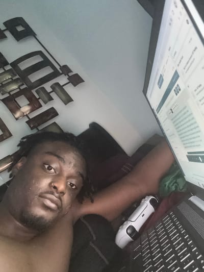 A close-up selfie-style photograph, taken from an overhead perspective, depicts a person lying down in an indoor setting in Arlington, United States of America. The subject is a male with dark skin, short dark hair styled in dreadlocks or twists, and a beard. He is shirtless and wears a small silver nose ring, looking directly at the camera with a neutral, calm expression.

Behind and to the left of the person's head, a piece of modern wall art is visible, consisting of various metallic, rectangular, and square shapes arranged geometrically against a light-colored wall. Above and to the right of the person, a computer monitor is prominently displayed, showing a web page or document with blocks of blurred text and user interface elements. Next to the monitor, a black laptop keyboard and a white gaming controller, resembling a PlayStation DualSense, are visible. The person's arm extends towards this area, suggesting they are interacting with or are in proximity to these devices.

The scene conveys a relaxed and casual atmosphere, likely in a bedroom or living area, indicating either leisure activity or remote work from a comfortable, recumbent position.