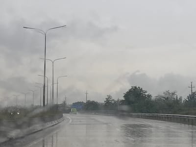 The image depicts a wet road with multiple streetlights and a green vehicle in the distance, captured in Etseri, Georgia. The sky is overcast with heavy clouds, suggesting rainy or foggy weather, which is further indicated by the water droplets on the camera lens, creating a blurred effect. The road appears to be a highway or a major road with a concrete barrier separating lanes and a grassy embankment on the left, lined with trees and more streetlights. A blue road sign is visible on the right side of the road, though its text is illegible due to the distance and atmospheric conditions. The overall scene conveys a sense of travel during inclement weather on a potentially remote or rural road.