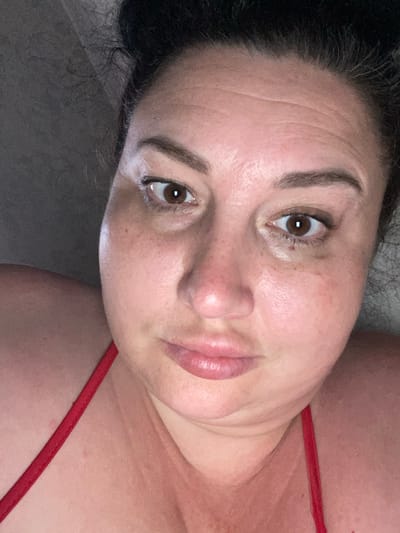 The image is a close-up selfie of a woman with dark hair pulled back. She is looking directly at the camera with a neutral expression. Her face is illuminated by natural light, highlighting her features, including her brown eyes, defined eyebrows, and slightly parted lips. She appears to be wearing a red garment, possibly a bikini top, as indicated by the strap visible on her left shoulder and across her chest. The background is dark and out of focus, suggesting an indoor setting. There are no discernible elements within the image that would allow for the identification of a specific location in Rozkvit, Ukraine.