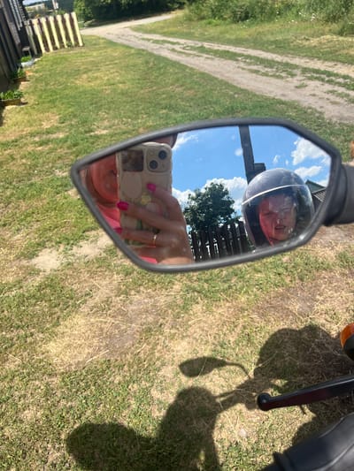 The image is a first-person perspective taken from a motorcycle, reflecting a rearview mirror. In the mirror, a person is visible holding a smartphone, taking a selfie. The person has bright pink fingernails and is wearing a pink top. To the right of the selfie, a child wearing a dark helmet with a clear visor is reflected, with their face visible through the visor. The child appears to be grimacing.

The background reflected in the mirror shows a dirt road with grass and trees on either side, suggesting a rural or suburban setting. There is a wooden fence and a building visible in the distance. The sky is blue with scattered white clouds. The foreground of the image, outside the mirror, shows the motorcycle's handlebars and a shadow cast on the dry, grassy ground, indicating a sunny day.

The location is identified as Сухринское, Russia.