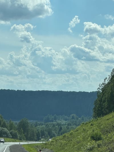 The image captures a panoramic view of a rural landscape under a sky filled with fluffy white clouds. In the foreground, a paved road curves to the left, with a white car visible on the left side of the road, moving away from the viewer. The road is bordered by vibrant green grass and bushes on the right side, and a sloping grassy hill with scattered vegetation on the far right.
In the mid-ground, a dense forest of green trees stretches across the scene, creating a textured carpet of foliage. Beyond the forest, a dark, undulating silhouette of a hill or mountain range forms the background. A faint body of water may be visible in the distance, nestled within the valley. The overall impression is one of a serene, natural environment, possibly captured during a drive through the countryside. There is no evidence to suggest the location is in the city of Челва, Russia.