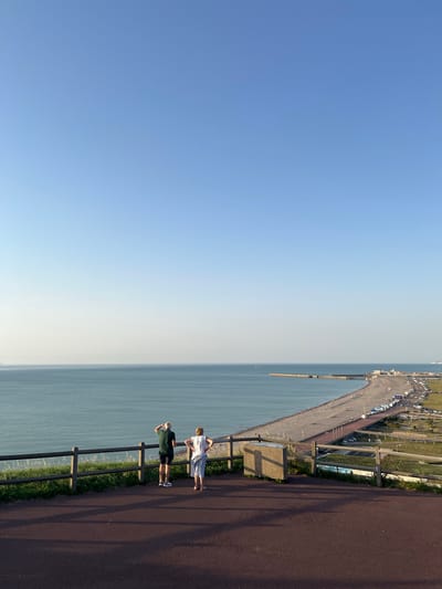 The image shows a view of the coast at Dieppe, France, taken from a high vantage point. The scene is dominated by a vast expanse of clear blue sky on the left, transitioning to a hazy, light-colored horizon where the sky meets the sea. The sea itself is a muted blue-green, stretching out to the horizon.
On the right side of the image, a paved pathway runs along the edge of a grassy area, bordered by a dark wooden fence. Two people are visible on the pathway, looking out towards the sea. One person is wearing a white dress, and the other is in a dark green t-shirt and dark shorts. They appear to be standing and observing the view.
Further along the coast, a long pier or breakwater extends into the sea, with several structures and vehicles visible along its length. A road with parked cars is also visible near the shoreline. The overall impression is of a scenic coastal location, possibly a promenade or a viewpoint.
The time of day appears to be late afternoon or early evening, given the long shadows cast by the fence and the warm lighting. The weather seems pleasant, with clear skies and calm seas. The location is identifiable as Dieppe, France. No visible text is present in the image.