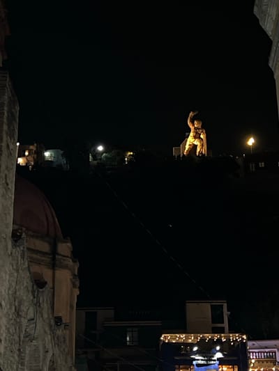 The image displays a nighttime urban scene in Guanajuato City, Mexico, centered on the illuminated El Pípila monument positioned atop a distant hill. The large statue, rendered in a golden hue by external lighting, depicts a male figure with a raised right arm holding an object resembling a torch or club. Below the monument, the hillside features scattered lights and subtle indications of other structures.

In the lower left foreground, a section of an aged stone building is visible, characterized by a weathered facade and a reddish-brown domed roof. Adjacent to this structure, extending into the midground, are various multi-story city buildings with flat roofs and illuminated windows. One such building in the lower right portion of the frame features a string of decorative white lights and a partially visible commercial sign that reads "VINYL BAR." A thin, dark diagonal wire traverses the central upper section of the image. The sky is dark and clear, without visible celestial bodies or atmospheric phenomena. No discernible human figures are present or engaged in specific actions.