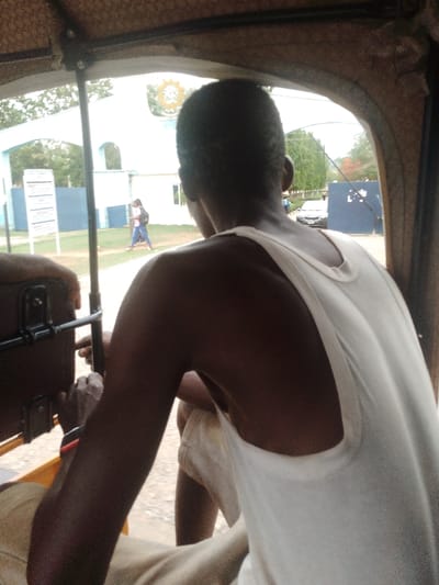 The image shows a young black man sitting in the back of a vehicle, presumably a bus, in the city of Abuja, Federal Capital Territory, Nigeria. The man is facing the back of the vehicle, wearing a plain white tank top and a pair of khaki shorts. His left arm is resting on the back of the seat in front of him, while his right arm is hanging down. The vehicle's window reveals a view of a building with a blue door and some greenery. There is another person visible outside the window, walking towards the vehicle. The scene appears to be during the day as the sun is shining. The environment suggests an urban setting with a building in the background and vegetation near the vehicle.  The coordinates provided suggest that the scene is in Abuja, the capital city of Nigeria.