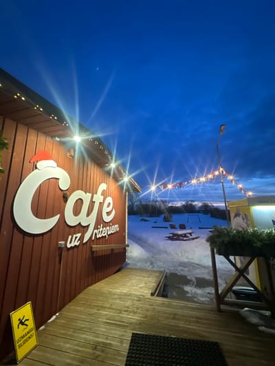 A sideways view of a cafe at dusk. The building, made of reddish-brown wood, is adorned with a large white "Cafe uz riteņiem" sign. Strings of lights run along the roof. A warning sign cautions of slippery surfaces. Beyond the cafe, a snow-covered area leads to a frozen body of water, lined with a string of holiday flags. The sky is a deep blue, with a sliver of moon visible.
