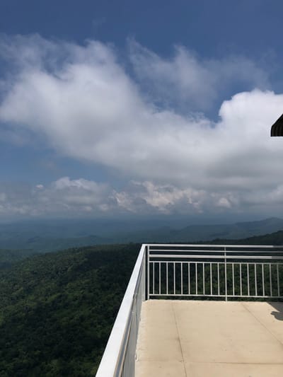 An elevated viewpoint overlooks a lush, green mountain range under a bright blue sky with scattered white clouds. A concrete balcony with a white metal railing occupies the foreground, suggesting a tourist spot or observation deck. The mountainous landscape stretches into the distance, with layers of hills fading into a hazy blue. No people are visible in the frame.