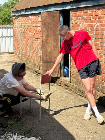 Two young men are in an outdoor area next to a brick building, possibly in Komsomolskiy, Russia, on a sunny day. The building has a rusty metal roof and a wooden door that is ajar. One man, wearing a white t-shirt, black pants with yellow stripes, and a welding mask, is crouching and working on a red-seated metal chair. He is wearing gloves and appears to be using a tool on the chair. The other man, wearing a red t-shirt with "USPA" printed on it, black shorts, white socks, and white clogs, is standing and leaning towards the chair, looking down. He has his hand on the back of the chair. In the background, there is a white corrugated metal fence and some greenery. The ground is paved with asphalt. There are electrical cords and wires on the ground near the crouching man.