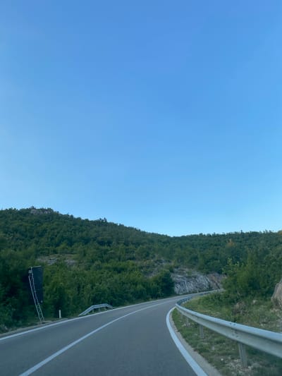The image shows a winding road passing through a hilly, forested area. The road is paved and has white lines marking the lanes. A metal guardrail runs along the right side of the road. The surrounding hills are covered in dense green trees. The sky is clear and blue. In the lower left portion of the image, a dark rectangular sign is visible, supported by a metal frame. The location is identified as Miločani, Montenegro. There are no people visible in the image.