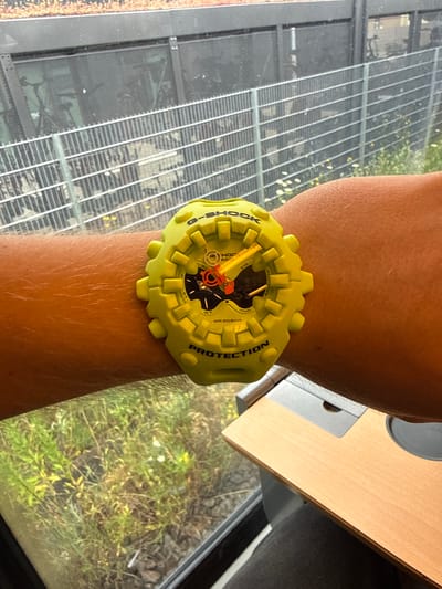 A bright yellow G-Shock watch is worn on a person's arm. The watch has an analog display with orange and black hands and a black face with white markings. The word "G-SHOCK" is visible on the watch face and bezel. The background shows a metal fence, green vegetation, and a wooden surface, possibly a desk. This scene is located in Bitterfeld-Wolfen, Germany.