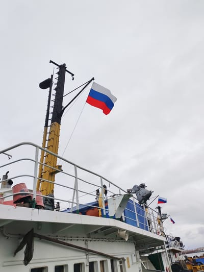 The image shows the upper deck of a vessel, likely a boat or ship, with multiple Russian flags displayed. A prominent flag flies from a tall mast, with two smaller flags visible on the structure behind the deck railing. The vessel appears to be stationary, with parts of its structure and equipment visible, including a light fixture, various railings, and what seems to be some sort of equipment housing in blue and white. The sky is overcast. Based on the presence of Cyrillic script on the vessel and the flags, this scene is likely in Russia, possibly Rybinsk. The image captures the upper portion of the vessel and the overcast sky above.
