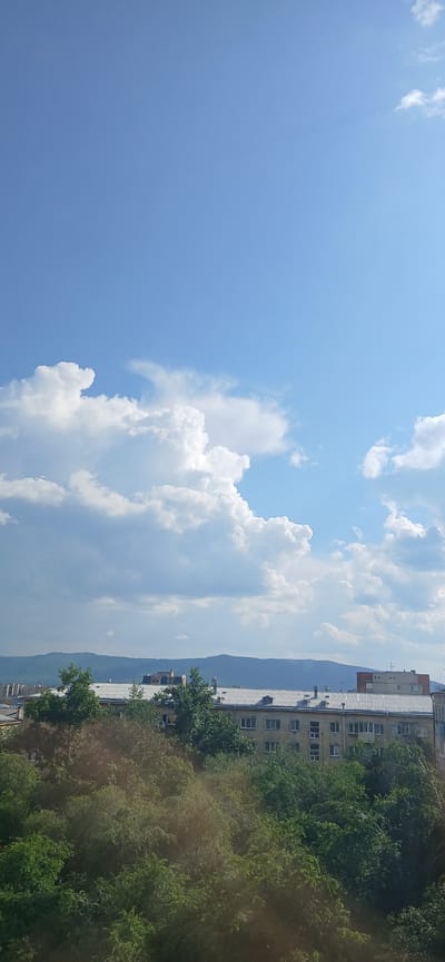A panoramic view of the city of Chita, Russia, is captured under a bright blue sky dotted with fluffy white clouds. In the foreground, lush green trees create a dense canopy. Behind the trees, a long, multi-story building with a white roof is visible, featuring numerous windows. Further in the distance, rolling hills form a backdrop to the urban landscape. The image appears to be taken during daylight hours, likely on a clear, sunny day.