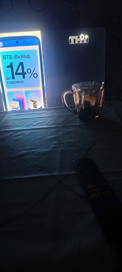 The image shows a close-up of a television screen displaying an advertisement for a VTB bank deposit, with a 14% annual interest rate. The screen is on a dark background, and a glass mug with a handle sits to the right of the screen. A black remote control is visible in the lower right corner of the frame. The overall scene appears to be indoors, with the lighting suggesting it is nighttime or the room is dark. The location is not specified as Хохлы, Russia.