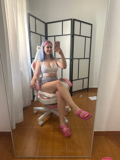 The image shows a young woman with blue and pink hair sitting in a white and pink gaming chair, taking a selfie in a full-length mirror. She is wearing a white crop top, grey shorts with a pattern, and pink slide sandals. The room has wooden floors and a large mirror. Behind her, a foldable room divider with white panels and a black frame is visible. A white curtain hangs to the left. The scene appears to be in a residential setting. No specific landmarks or indicators place this image within the city of Oviedo/uviéu, Spain.