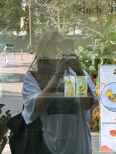 A reflection in a window shows a woman taking a photo with her phone. The reflection is overlaid with the restaurant's menu displaying pasta dishes. Outside, a street with trees and a Tipbox advertisement is visible. The woman wears a white t-shirt, and her hair is blonde. The restaurant's interior is hinted at with leafy plants.
