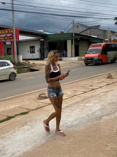 This media file shows a woman standing on a sidewalk next to a road, holding a smartphone. She has blonde hair and is wearing a black crop top, denim shorts, and pink flip-flops. She has visible tattoos on her arms and legs.
In the background, there is a red vehicle, possibly a bus or van, parked on the side of the road. There are also several buildings visible, some with a modern architectural style and others that appear to be older. The scene suggests an outdoor, potentially urban or suburban setting. The presence of power lines and street signs indicates a populated area.
The weather appears to be overcast, with dark clouds visible in the sky. The road is paved, and the sidewalk is made of concrete. There are some plants and debris on the side of the road.
The text visible in the image, in Vietnamese, appears to be signage for a "HÁCH SA HOẠT THỦY PHỤC VỤ 24/24", which translates to "Hach Sa Operation Water Service 24/24". Another sign reads "CON PHONG". The location context provided is Thôn 2, Vietnam. The woman's expression is neutral as she looks at her phone. The overall impression is a snapshot of daily life in Vietnam.
