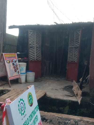 The image shows a weathered, open-fronted establishment in Iju, Nigeria. The structure is made of corrugated metal roofing and dark, possibly tarp-like, material for the walls. Two wooden doors, with louvered panels, are partially open, revealing the dark interior which is draped with vertical strands of material, possibly plastic or fabric. In front of the establishment, a sandwich board sign displays menu items for "COMFORT RESTAURANT CALABAR KITCHEN (ALL KINDS OF LOCAL FOOD)". The sign also includes images of various dishes and a phone number. Beside the sign, three white plastic buckets are stacked, one of which has "TOP BOND" written on it. The ground in front of the establishment is concrete and appears wet and somewhat dirty. To the left of the main structure, another sign, partially visible, has a green gear logo and text that includes "IEAKO-LIHYE LOCAL GOVERNMENT". The overall environment suggests an outdoor, possibly commercial setting in a developing area, with elements of makeshift construction.